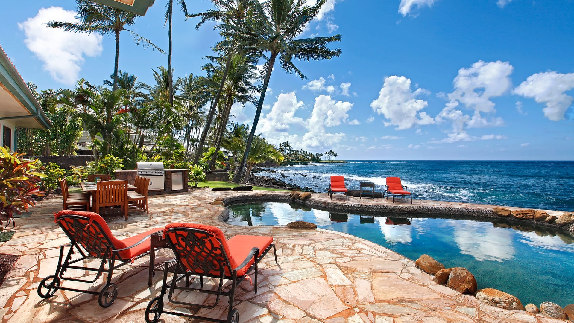 A tropical backyard leads out to the ocean, with a stone patio, a cozy pool, and bright orange loungers facing the waves. It’s the kind of space made for grilling, relaxing, and enjoying the sound of the water all day long.