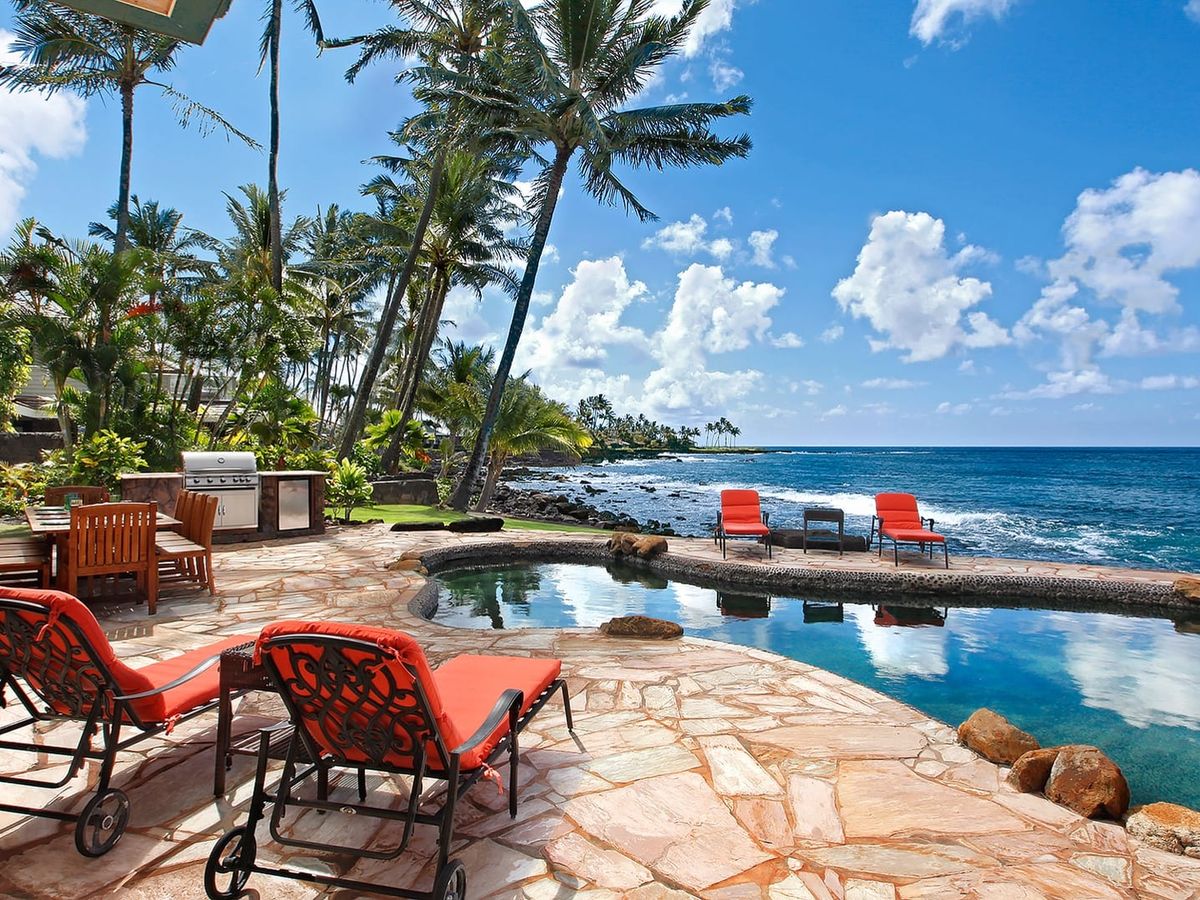 A tropical backyard leads out to the ocean, with a stone patio, a cozy pool, and bright orange loungers facing the waves. It’s the kind of space made for grilling, relaxing, and enjoying the sound of the water all day long.