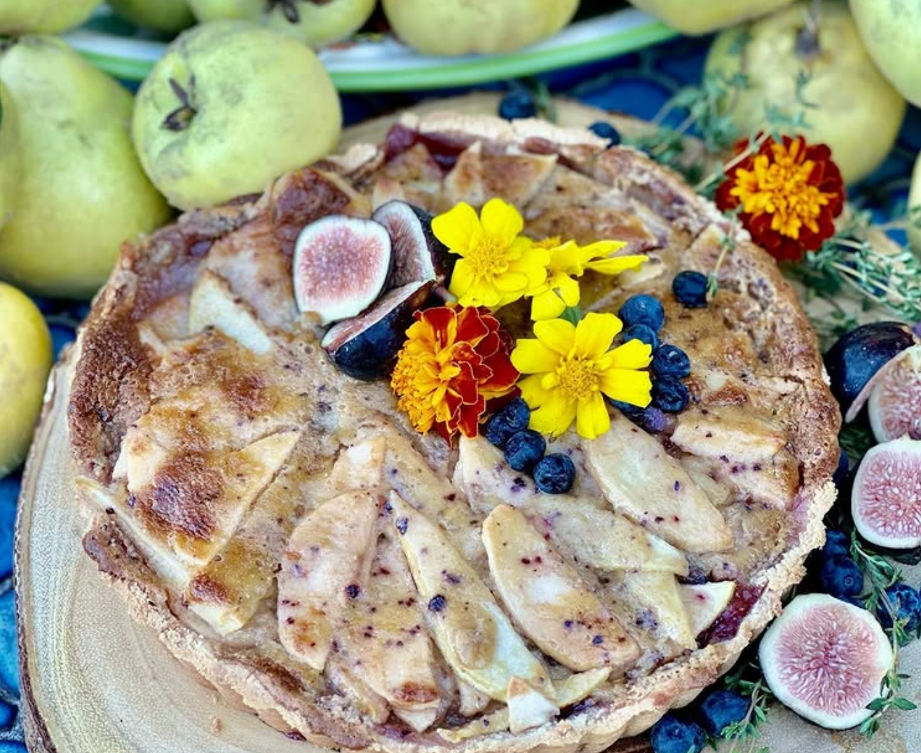 A rustic fruit tart topped with sliced apples, fresh figs, blueberries, and edible flowers, surrounded by pears and figs.