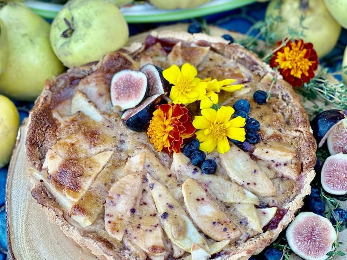 A rustic fruit tart topped with sliced apples, fresh figs, blueberries, and edible flowers, surrounded by pears and figs.