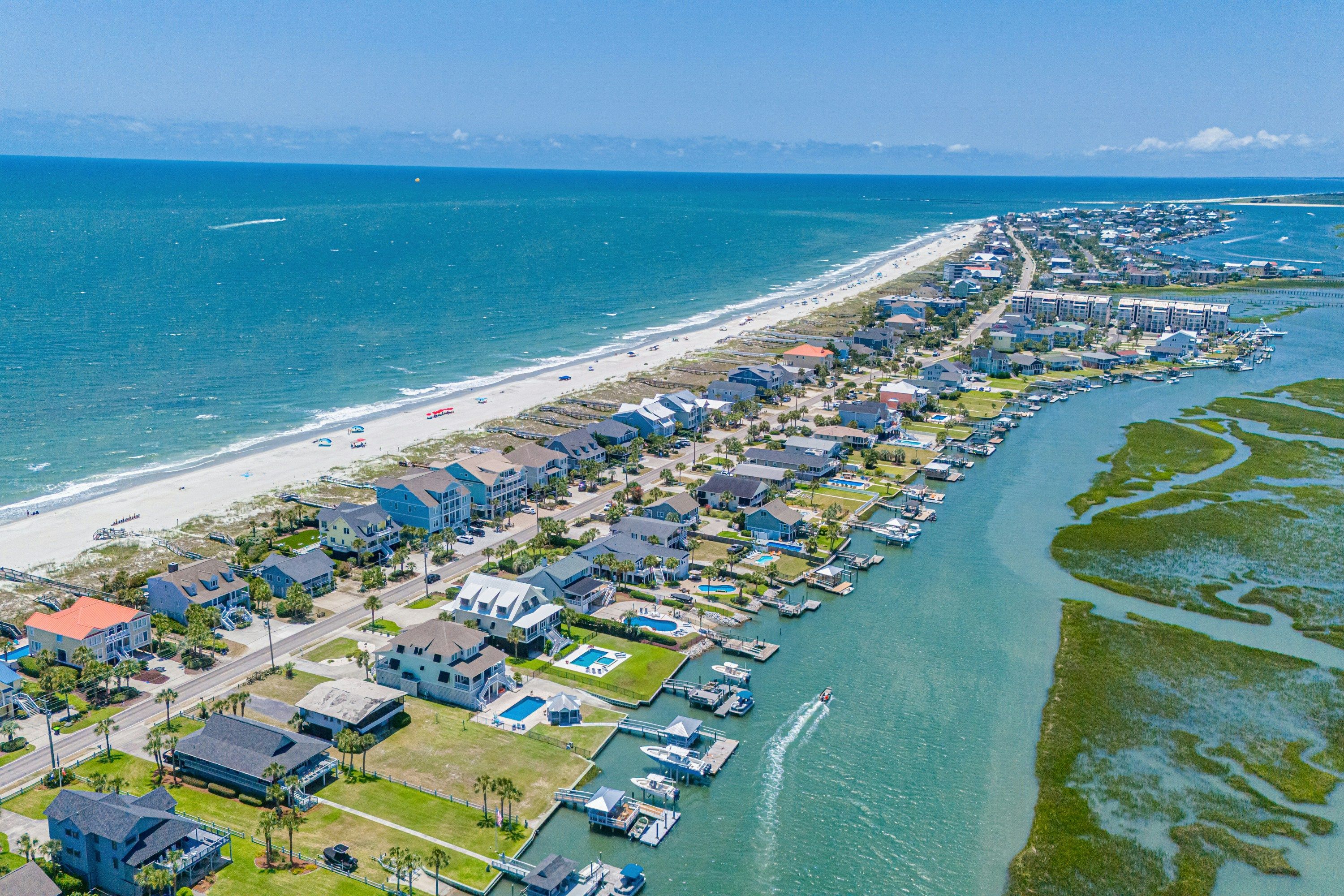 An aerial view of a coastal neighborhood in the Myrtle Beach area, with oceanfront homes lining a sandy beach on one side and calm marsh waterways with docks and boats on the other under a clear blue sky.
