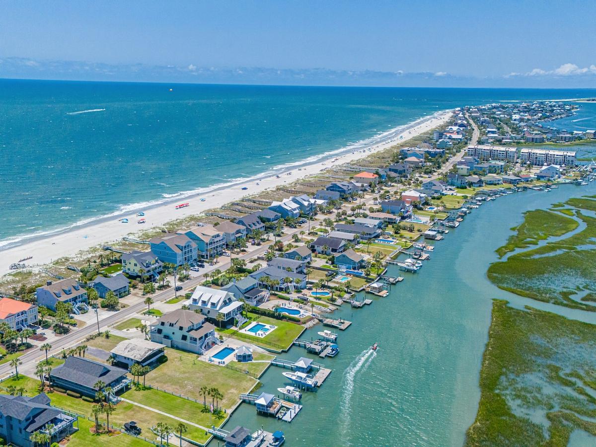 An aerial view of a coastal neighborhood in the Myrtle Beach area, with oceanfront homes lining a sandy beach on one side and calm marsh waterways with docks and boats on the other under a clear blue sky.