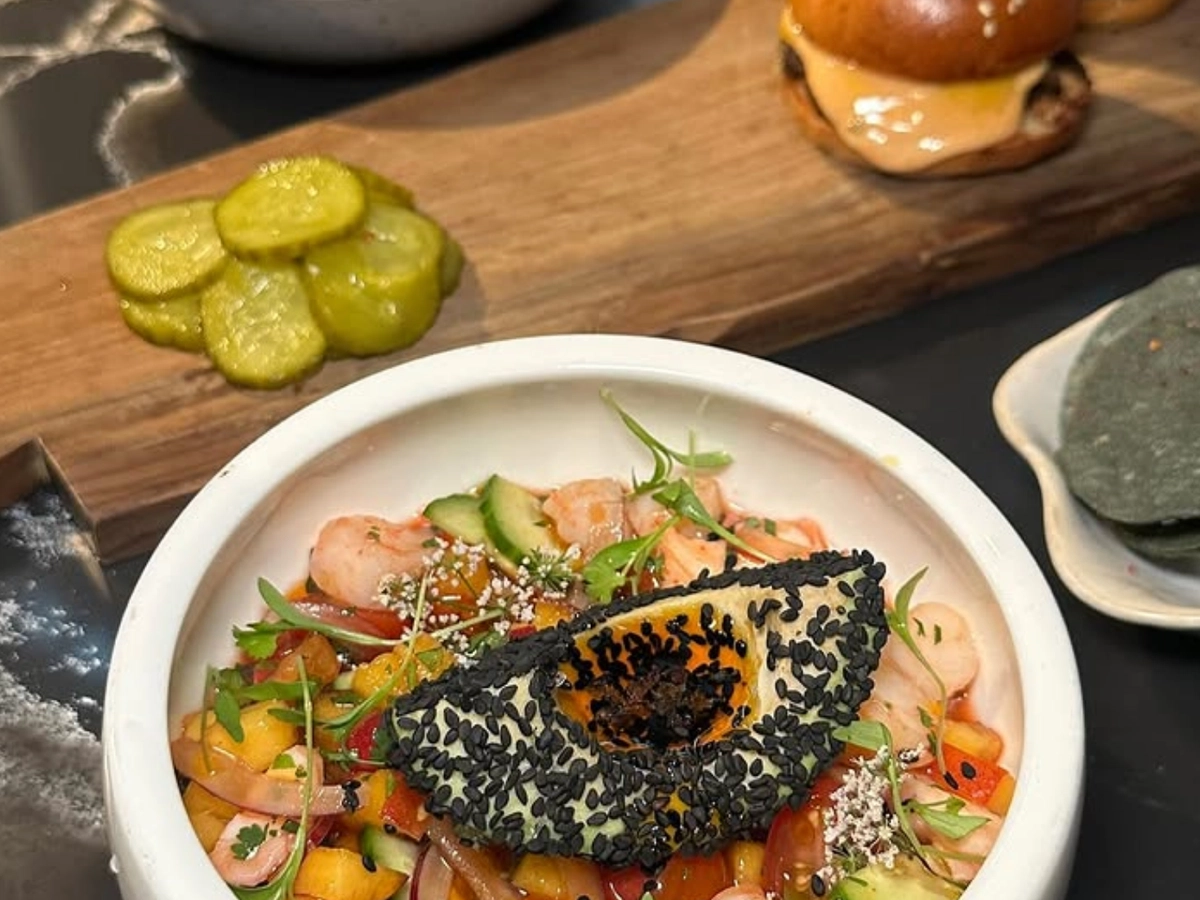 Colorful bowl of ceviche topped with a sesame-crusted avocado slice, served alongside sliders, croquettes, and pickle slices on a wooden board.