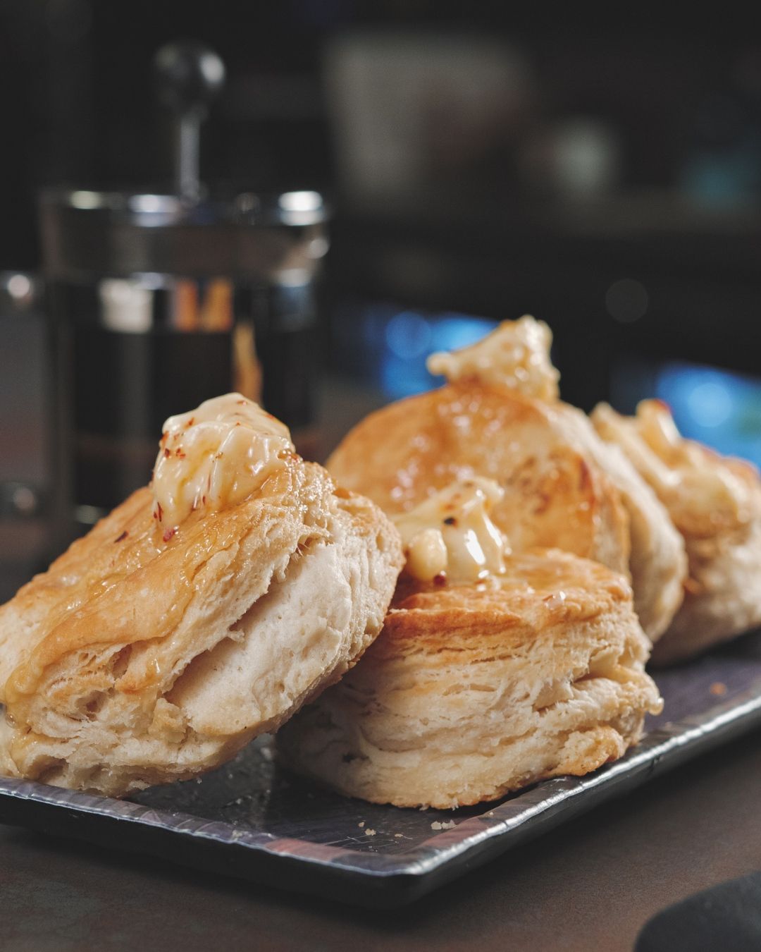 Two golden, flaky biscuits rest on a dark slate plate, split open and topped with melting butter and a drizzle of honey. The soft, layered interior of the biscuits is visible in the foreground, with a blurred salt shaker and bokeh lights in the background. 