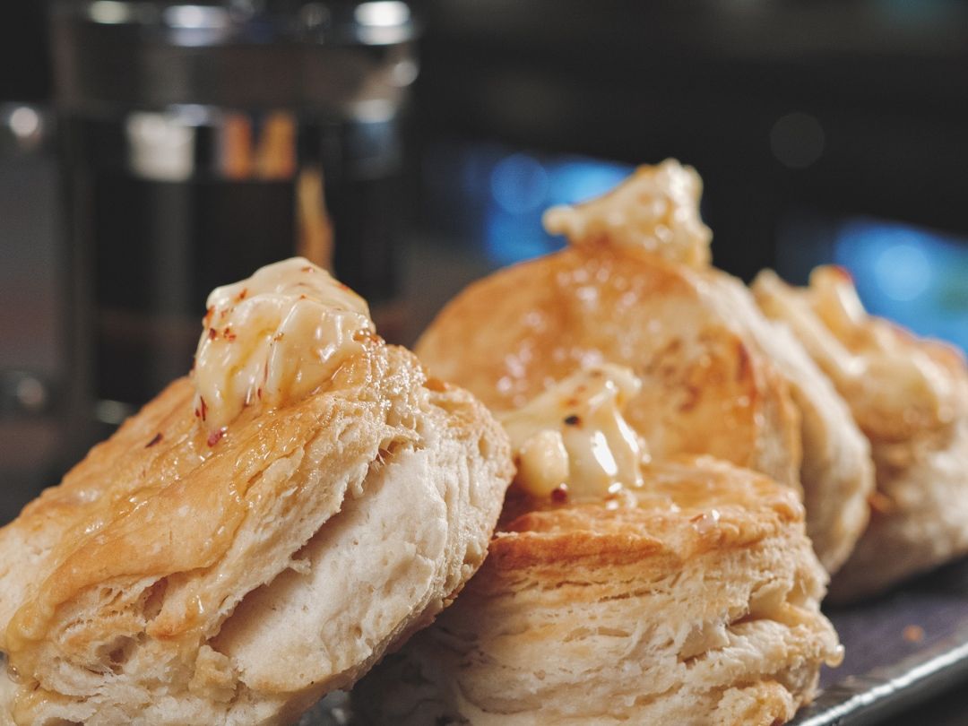 Two golden, flaky biscuits rest on a dark slate plate, split open and topped with melting butter and a drizzle of honey. The soft, layered interior of the biscuits is visible in the foreground, with a blurred salt shaker and bokeh lights in the background.