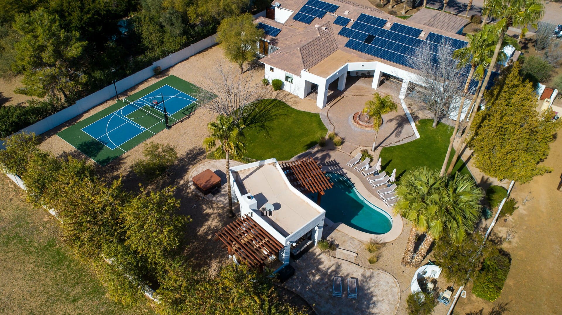 Aerial view of a solar-powered desert estate with a basketball court, resort-style pool, and lush landscaping.