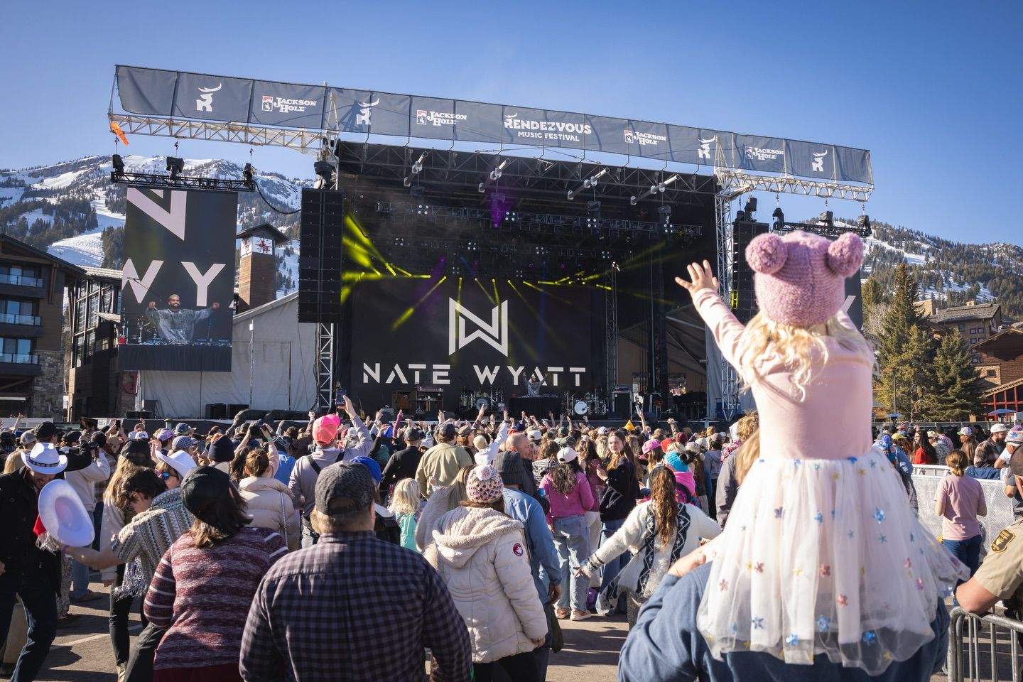 A large crowd gathers at the Rendezvous Music Festival with mountains in the background and live music on stage. Families and fans enjoy the outdoor concert atmosphere in downtown Jackson Hole. This annual event brings top artists to the area.