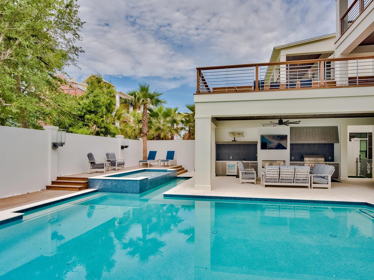 A stylish outdoor pool area features a raised spa, multiple lounge chairs, and a covered patio with a TV, grill, and dining space, surrounded by palm trees and a privacy wall.
