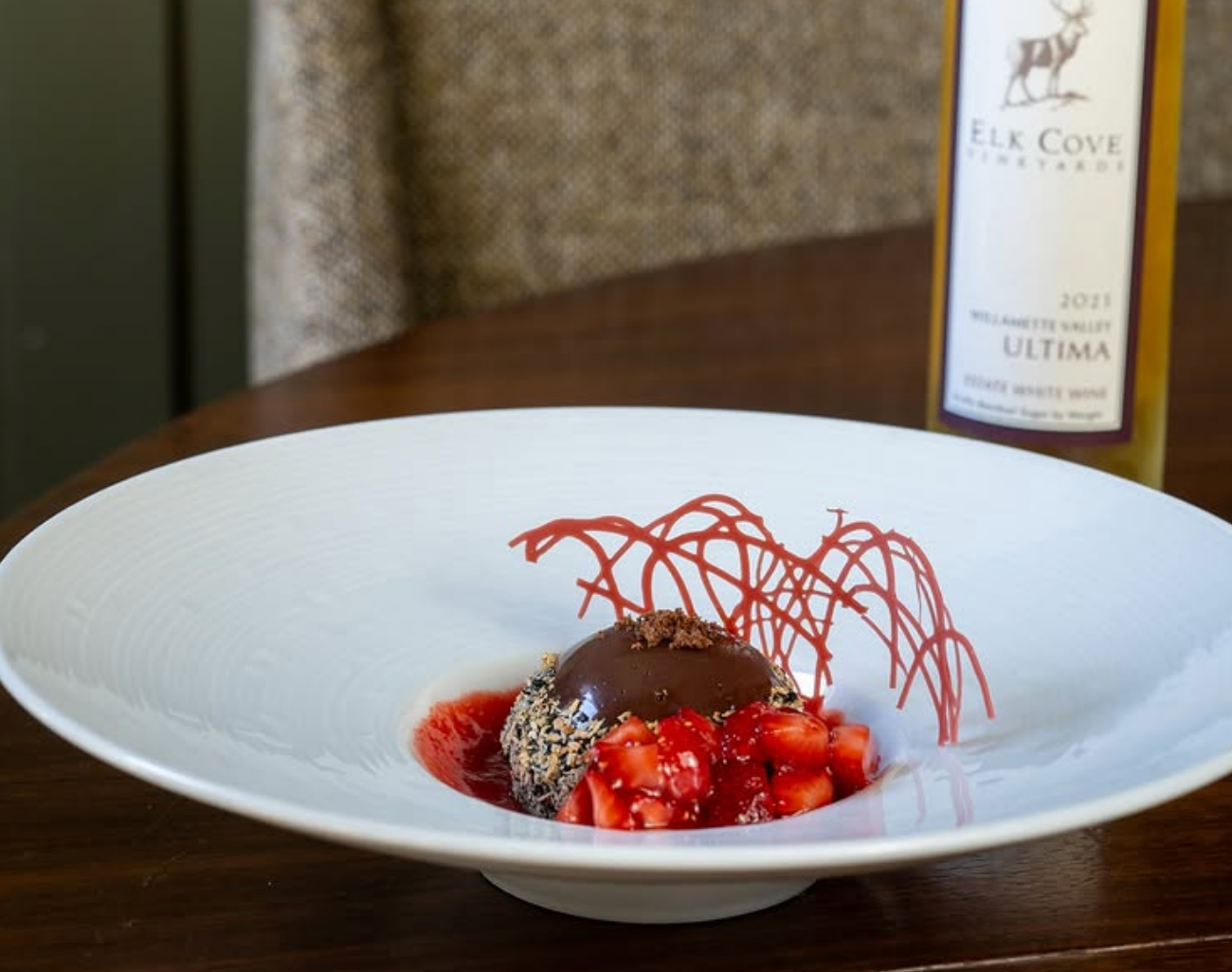 A plated chocolate dessert with fresh strawberries, red sauce, and delicate sugar garnish, paired with a bottle of Elk Cove Vineyards wine in the background.