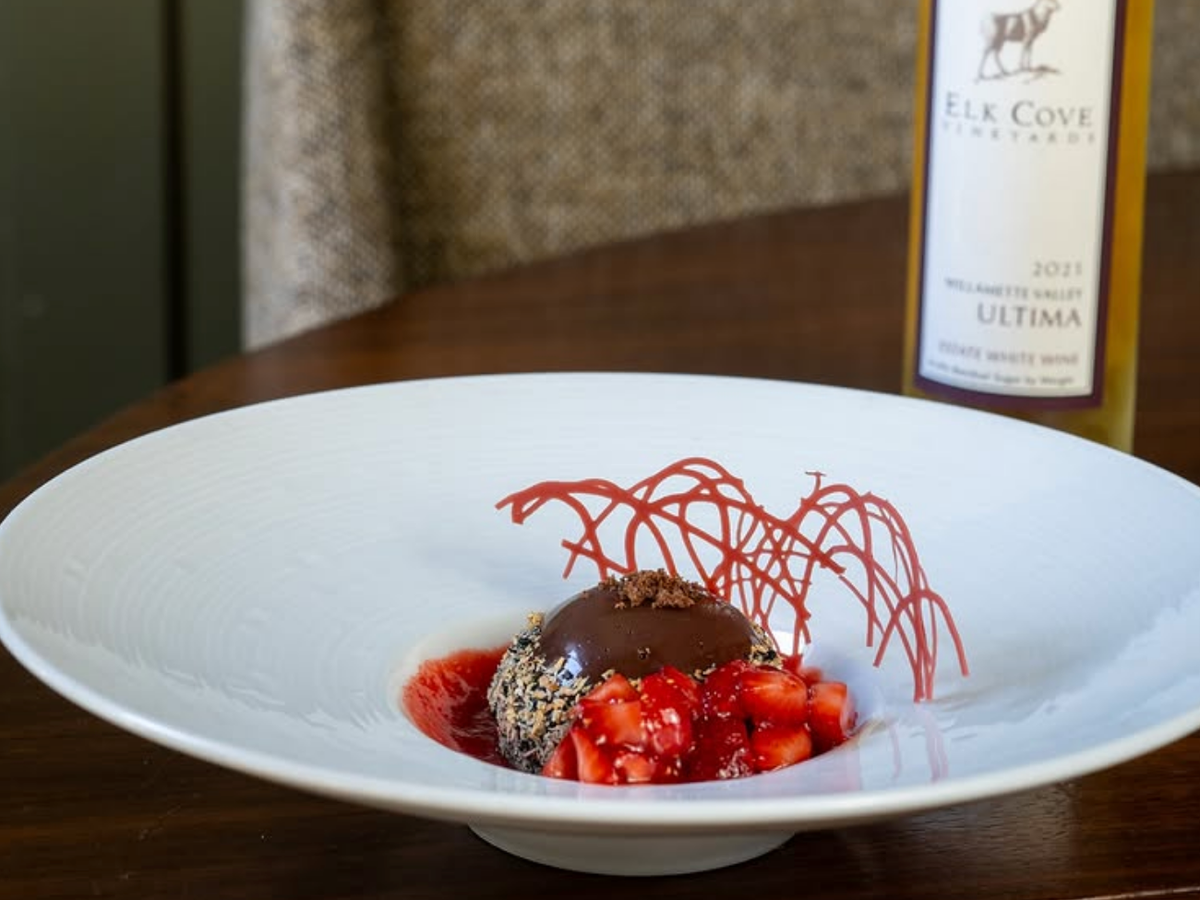 A plated chocolate dessert with fresh strawberries, red sauce, and delicate sugar garnish, paired with a bottle of Elk Cove Vineyards wine in the background.