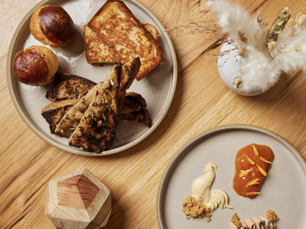 Overhead view of a wooden table set with two ceramic plates—one with assorted breads and the other with artistic swirls of sauces and garnishes—next to a geometric wooden container and a vase of dried florals.