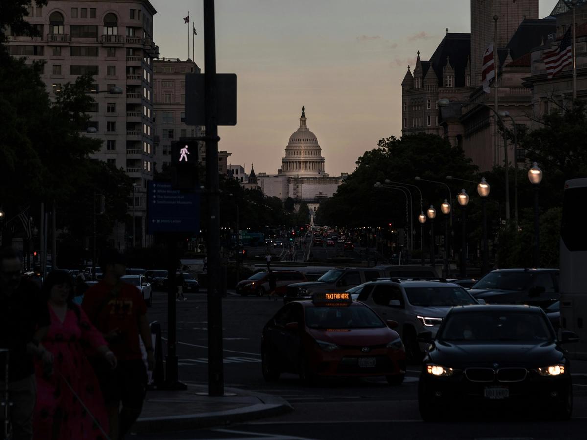Evening street view of Washington, D.C. with traffic, pedestrians, and the U.S. Capitol building illuminated in the distance against a dusky sky.