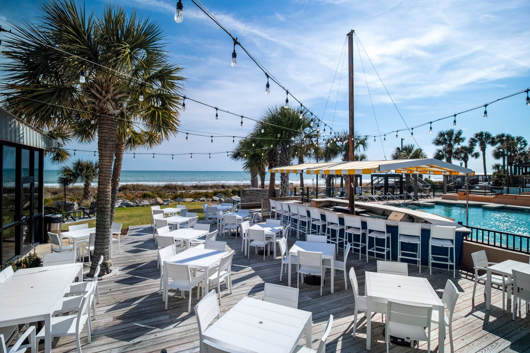 The oceanfront deck at Sea Captain's House in Myrtle Beach, South Carolina, features white outdoor dining tables and chairs strung with Edison bulb lights, a poolside bar with a yellow and white striped canopy, and sweeping views of the Atlantic Ocean framed by swaying palm trees. 