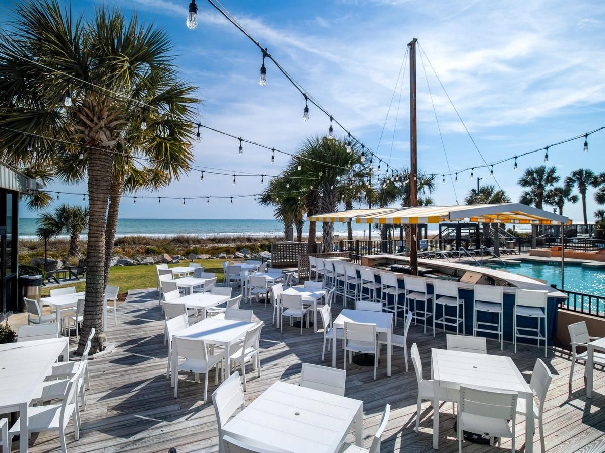 The oceanfront deck at Sea Captain's House in Myrtle Beach, South Carolina, features white outdoor dining tables and chairs strung with Edison bulb lights, a poolside bar with a yellow and white striped canopy, and sweeping views of the Atlantic Ocean framed by swaying palm trees.