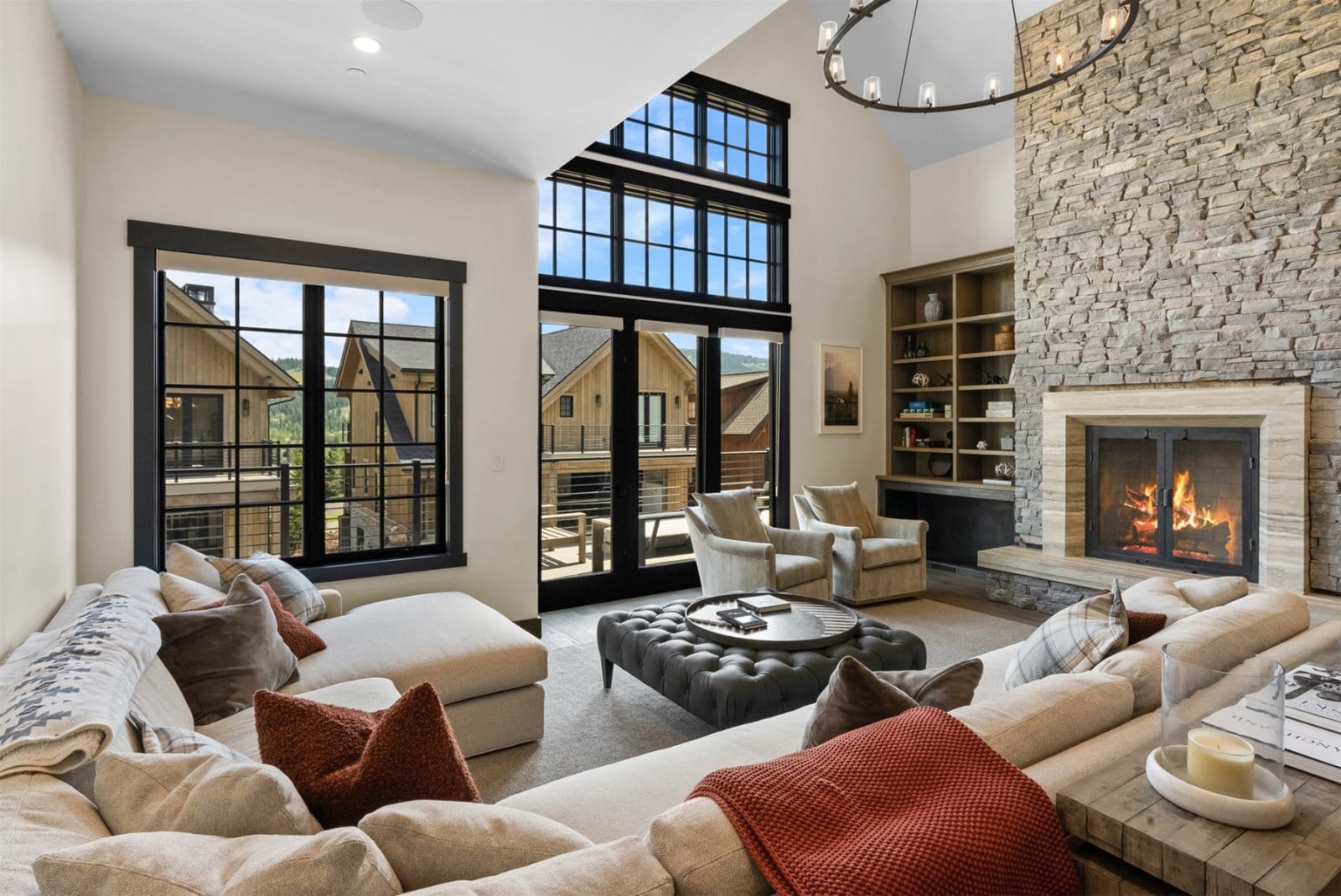 A modern mountain living room with a stone fireplace, cozy sectional sofa, and large windows offering views of neighboring cabins and the surrounding landscape.