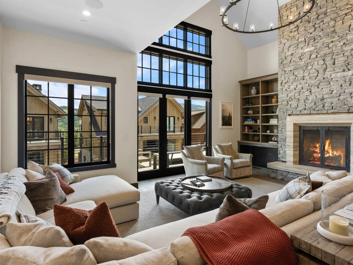 A modern mountain living room with a stone fireplace, cozy sectional sofa, and large windows offering views of neighboring cabins and the surrounding landscape.