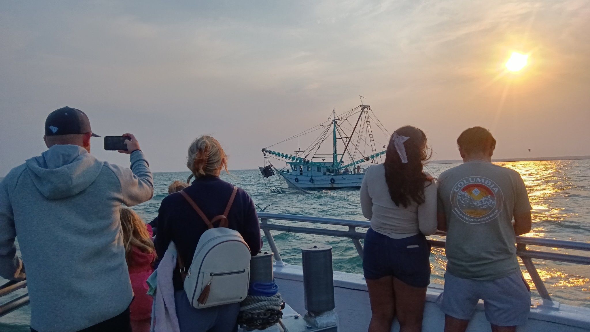 Visitors stand on a boat at sunset while watching a fishing trawler move across the water on a Myrtle Beach dolphin cruise. This photo captures the laid-back coastal feel of deep sea fishing and dolphin tours in Myrtle Beach, with golden light reflecting off the ocean.