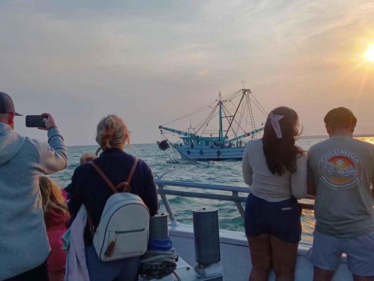Visitors stand on a boat at sunset while watching a fishing trawler move across the water on a Myrtle Beach dolphin cruise. This photo captures the laid-back coastal feel of deep sea fishing and dolphin tours in Myrtle Beach, with golden light reflecting off the ocean.