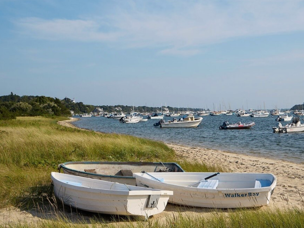 Two small white rowboats resting on grassy sand near a harbor filled with anchored boats under a clear blue sky