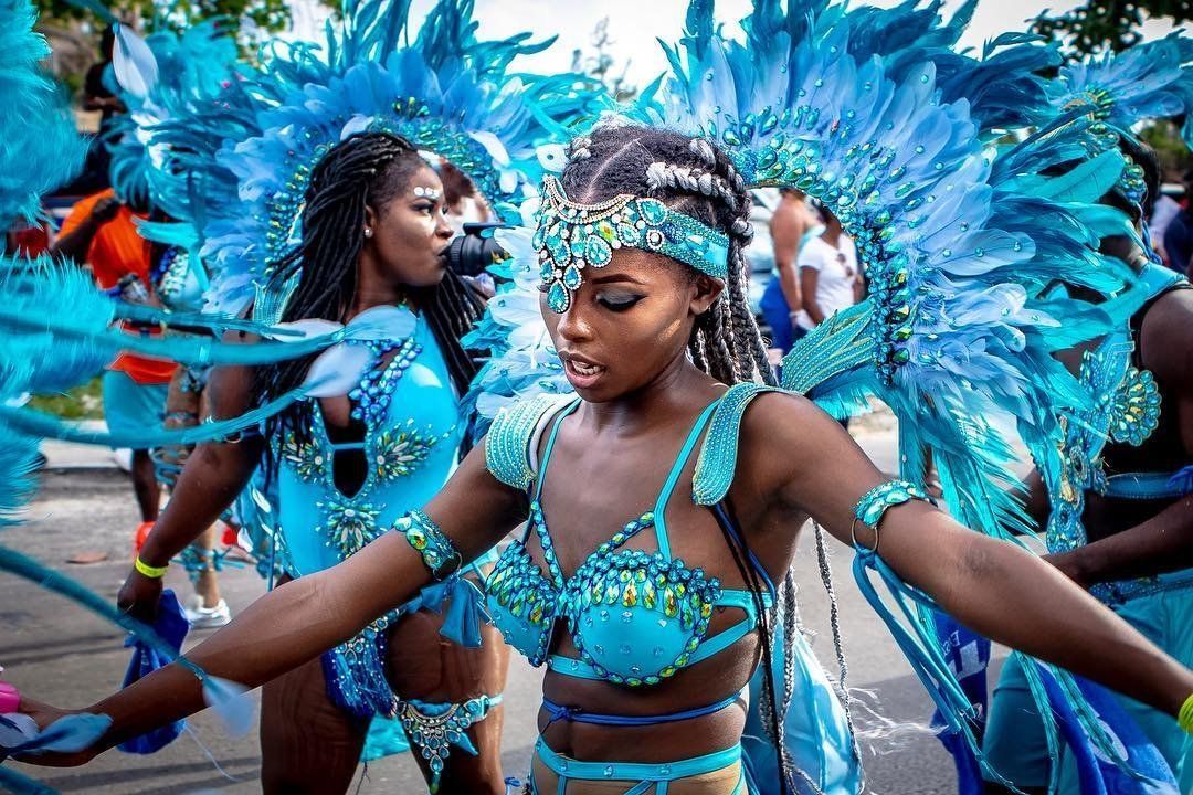 A vibrant Caribbean carnival dancer in a bright blue feathered costume moves through a street parade with arms outstretched. The detailed beads, feathers, and headpiece capture the energy, culture, and celebration of a traditional carnival festival. The scene feels lively, colorful, and full of movement during a cultural street event.