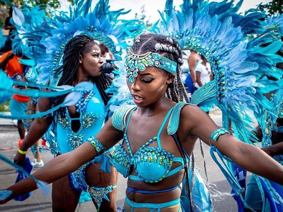 A vibrant Caribbean carnival dancer in a bright blue feathered costume moves through a street parade with arms outstretched. The detailed beads, feathers, and headpiece capture the energy, culture, and celebration of a traditional carnival festival. The scene feels lively, colorful, and full of movement during a cultural street event.