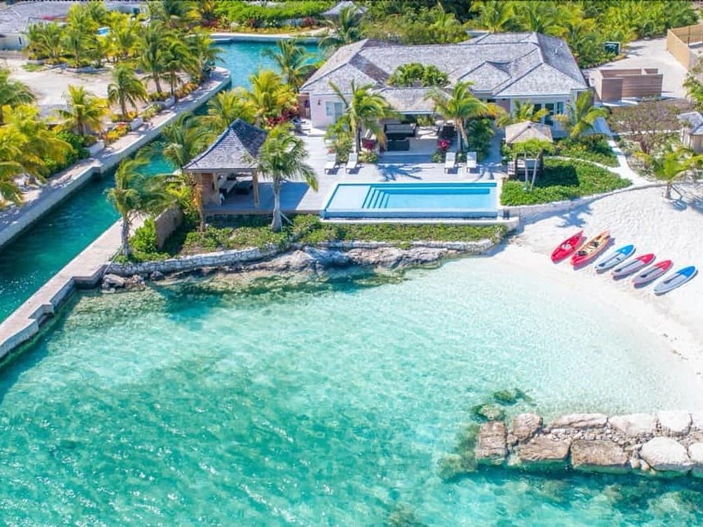 A luxury beachfront villa with a private pool and shaded cabanas overlooks turquoise waters, with colorful kayaks lined up on the white sand shore.