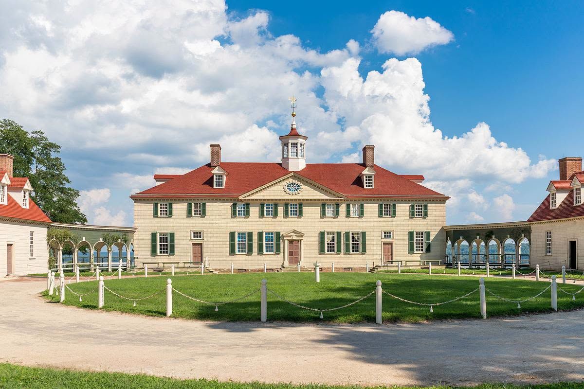 The iconic facade of George Washington's Mount Vernon estate in Virginia features its distinctive red roof, green shutters, and cupola with weathervane, flanked by colonnaded wings and a manicured lawn under a dramatic blue sky with billowing clouds. 