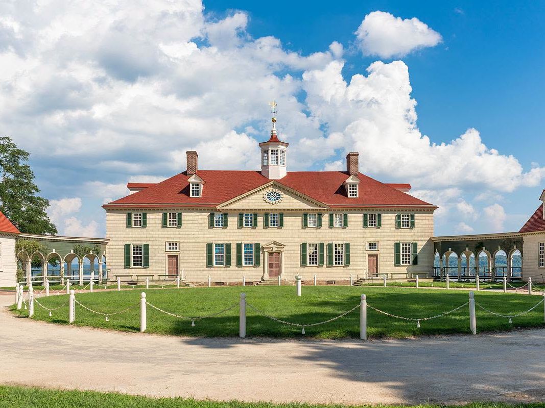 The iconic facade of George Washington's Mount Vernon estate in Virginia features its distinctive red roof, green shutters, and cupola with weathervane, flanked by colonnaded wings and a manicured lawn under a dramatic blue sky with billowing clouds.