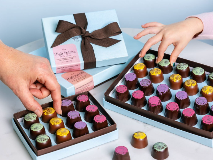 Hands reaching for colorful artisan chocolates in light blue gift boxes tied with brown ribbons.
