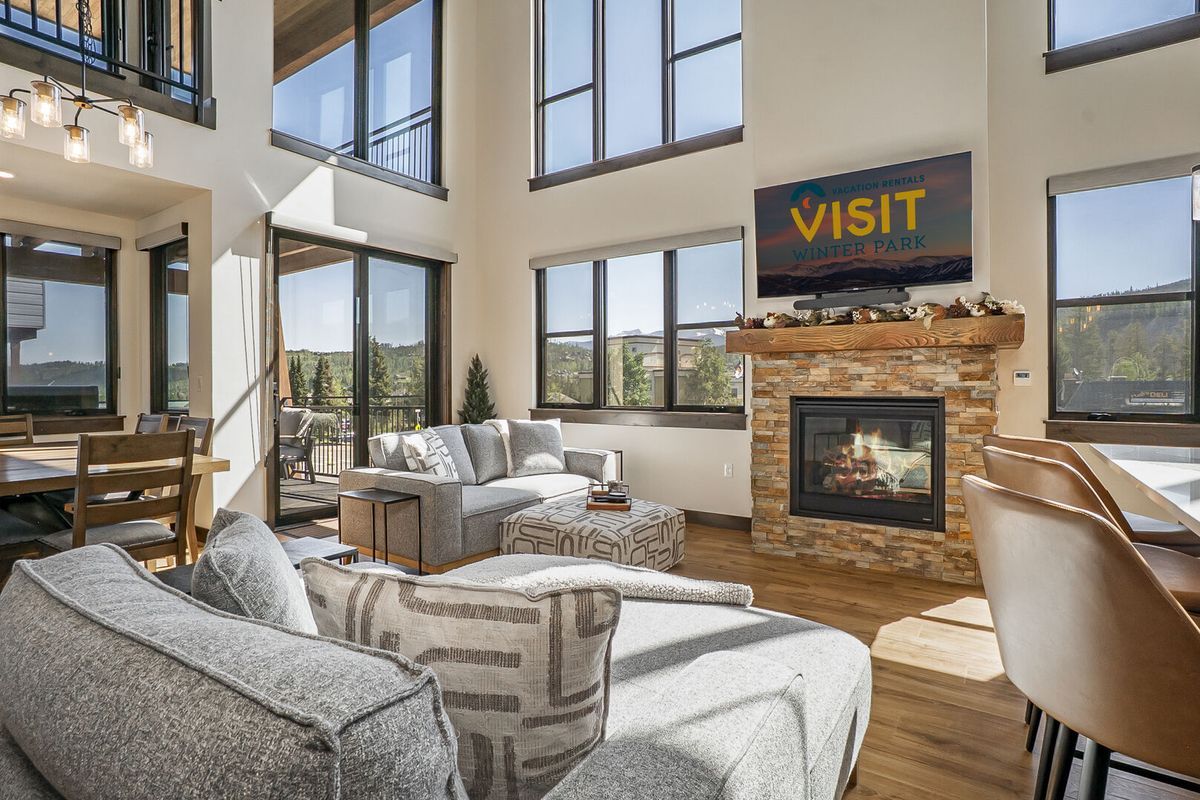 A bright and modern mountain living room featuring tall windows, a cozy gas fireplace with a stone surround, neutral-toned sectional sofas, and a flat-screen TV displaying “Visit Winter Park.”