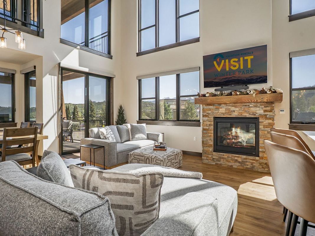 A bright and modern mountain living room featuring tall windows, a cozy gas fireplace with a stone surround, neutral-toned sectional sofas, and a flat-screen TV displaying “Visit Winter Park.”
