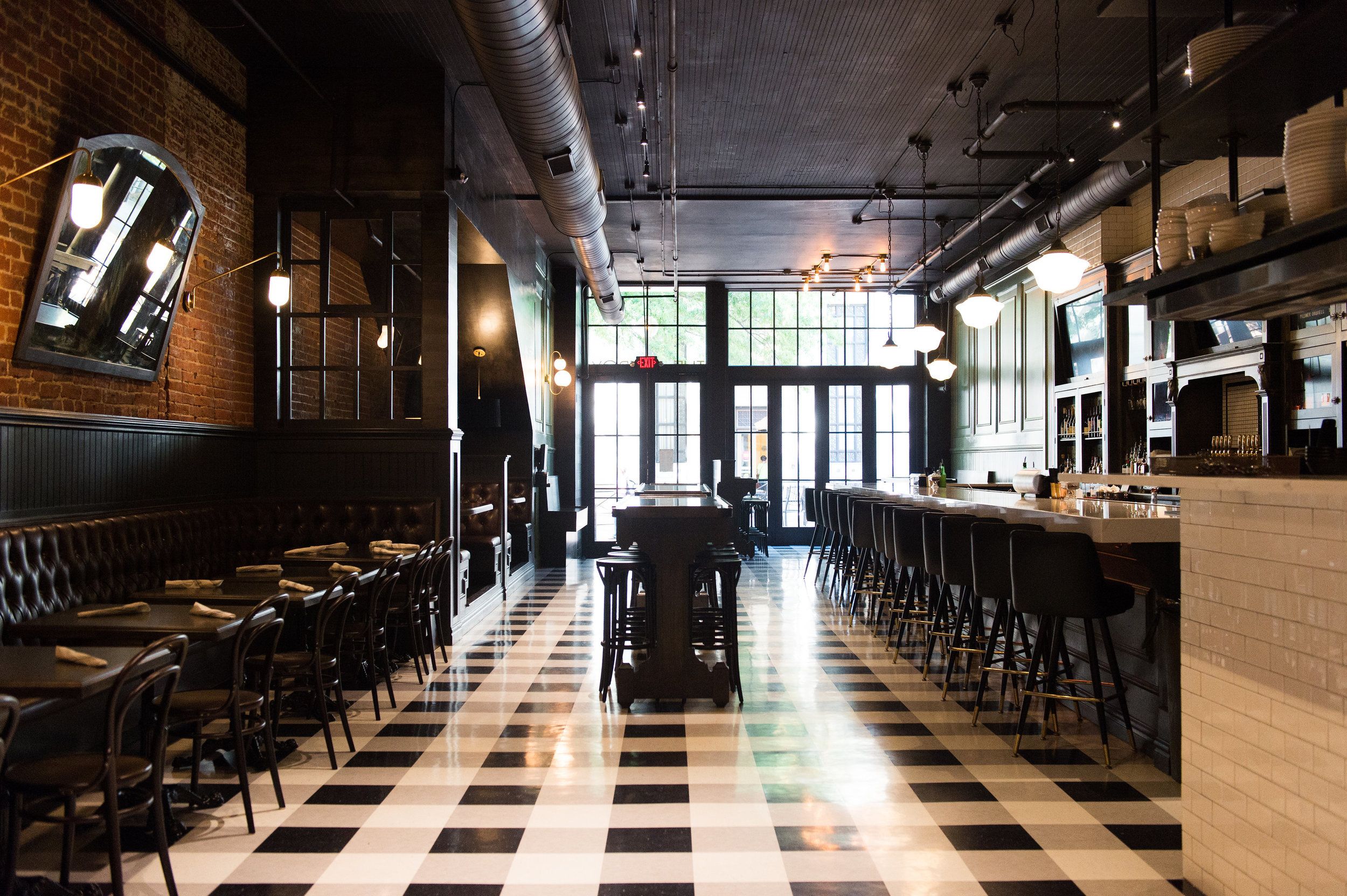 tzroy's dining room features a dramatic black-and-white checkered floor, exposed brick walls, and a long marble bar lined with dark barstools. Edison bulbs and globe pendants cast warm light throughout the industrial-chic space.