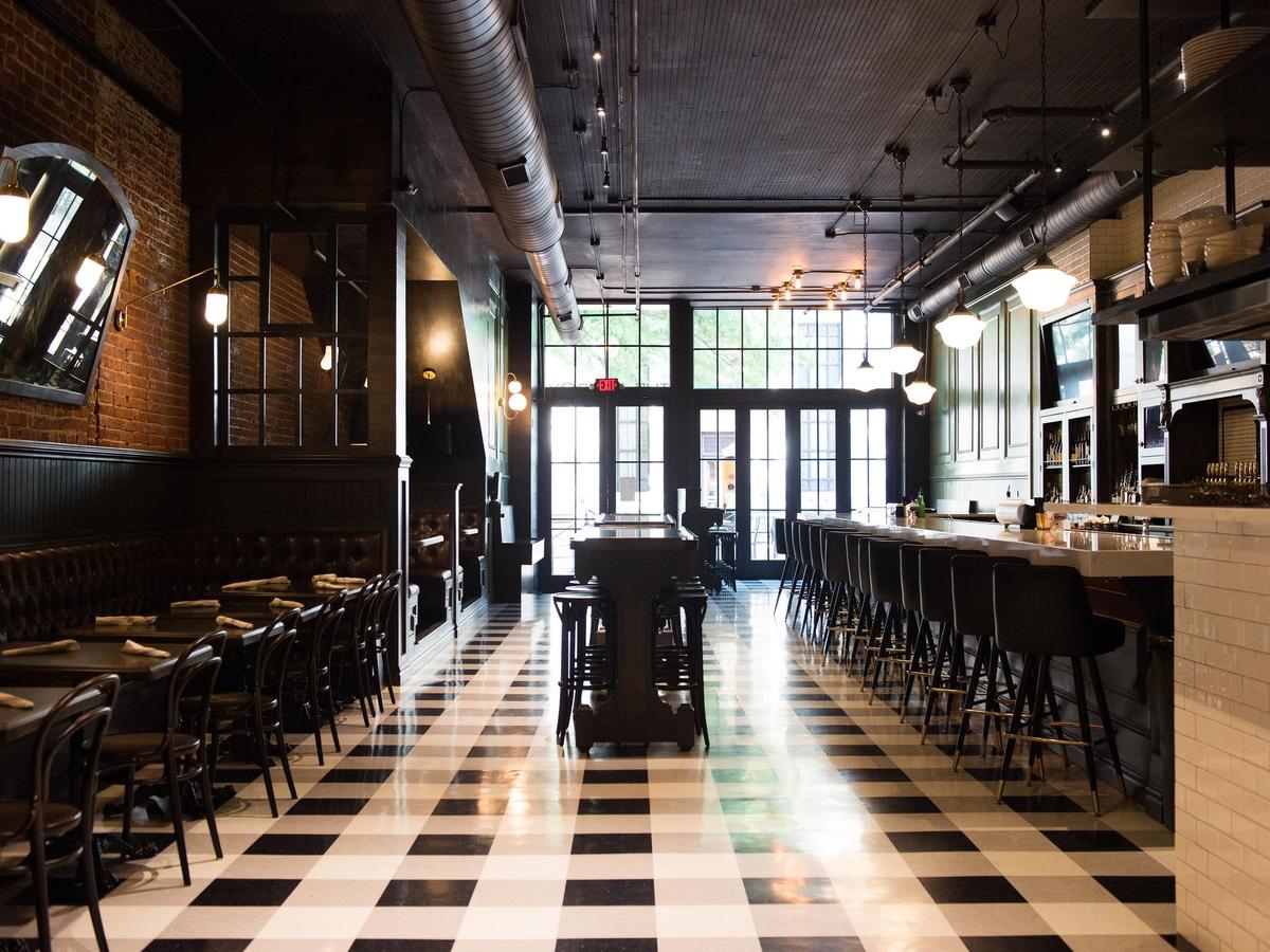 tzroy's dining room features a dramatic black-and-white checkered floor, exposed brick walls, and a long marble bar lined with dark barstools. Edison bulbs and globe pendants cast warm light throughout the industrial-chic space.