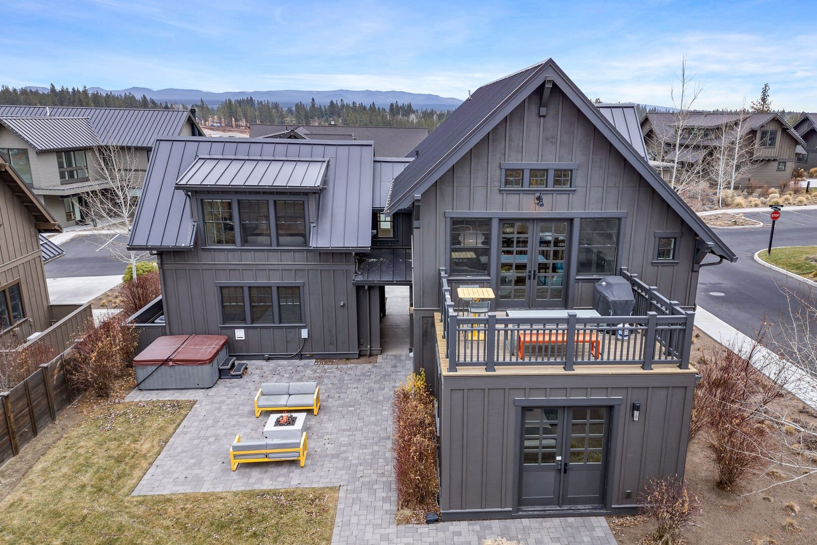 A modern mountain home with dark exterior siding, large windows, and a stylish outdoor patio complete with fire pit and lounge seating. The upper balcony and nearby hot tub make it a perfect blend of comfort and outdoor living.