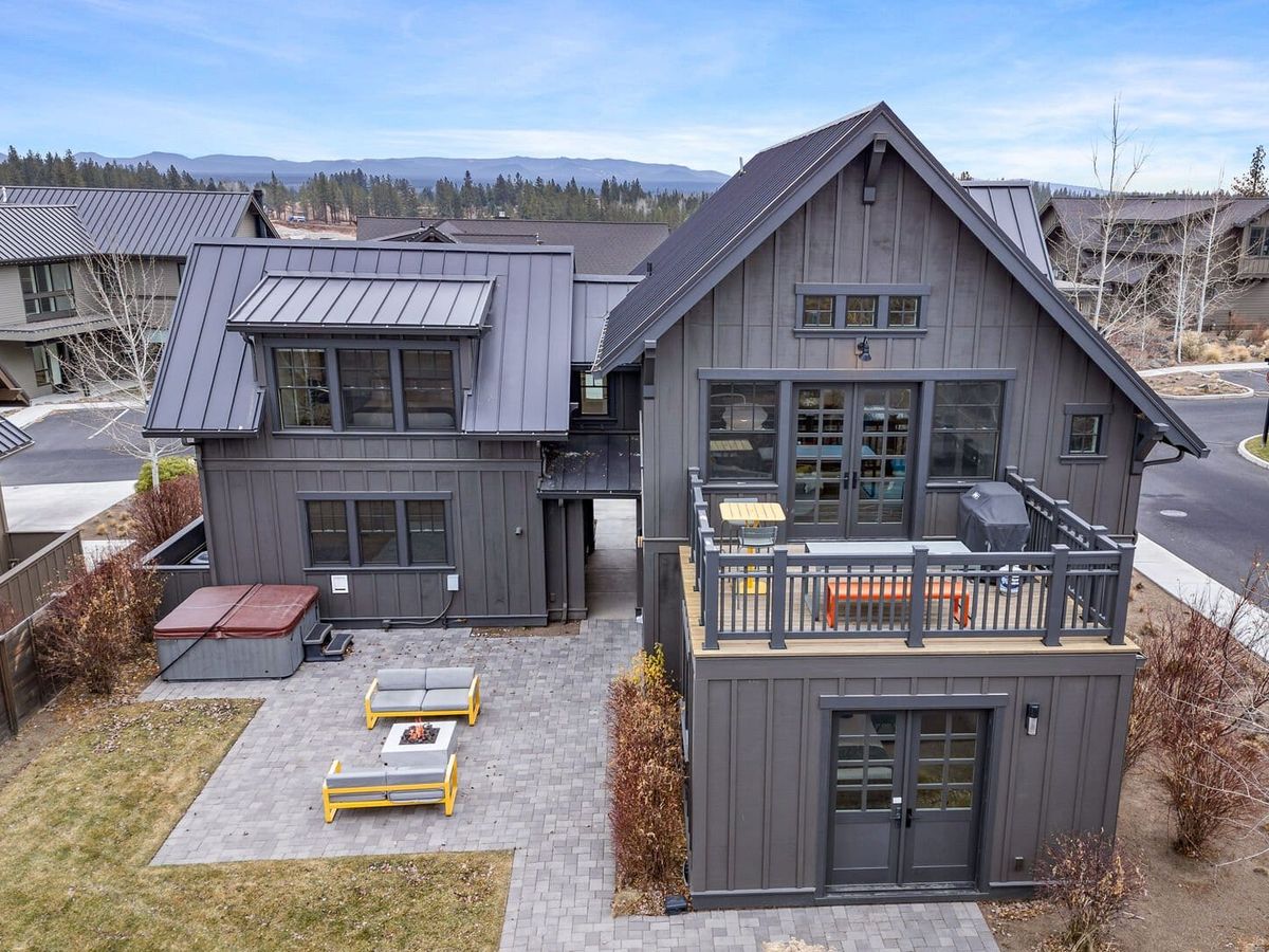 A modern mountain home with dark exterior siding, large windows, and a stylish outdoor patio complete with fire pit and lounge seating. The upper balcony and nearby hot tub make it a perfect blend of comfort and outdoor living.