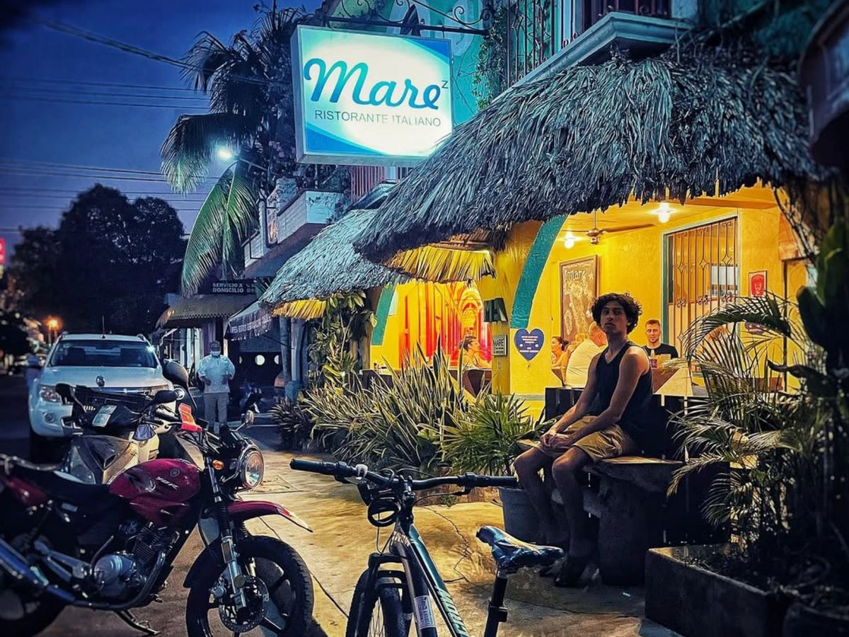 A vibrant evening street scene outside Maré Ristorante Italiano, with a thatched roof, warm yellow lighting, motorcycles parked out front, and people dining and relaxing on a bench under tropical foliage.
