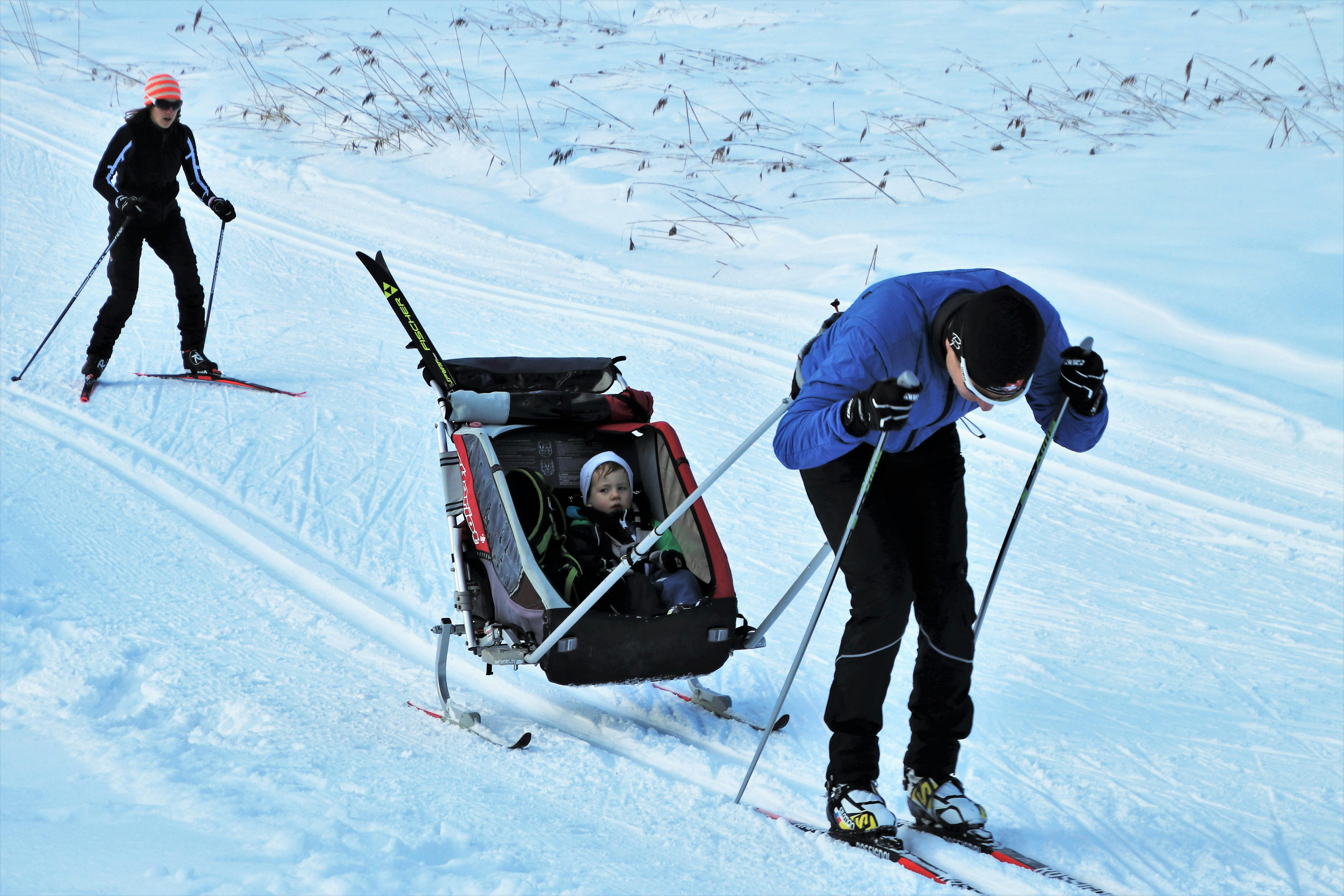 A parent cross-country skis along a snowy trail while pulling a child in a ski stroller. Another skier follows behind on the groomed winter path surrounded by open snow fields. This family-friendly winter scene shows how you can enjoy outdoor skiing even with young kids.
