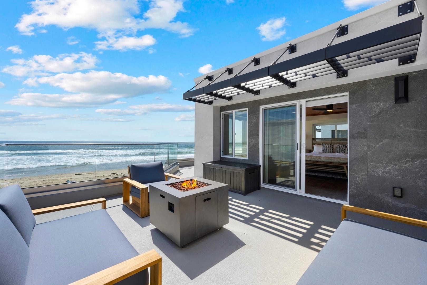 Oceanfront rooftop terrace with modern lounge seating, a sleek fire pit, and glass railing framing panoramic beach views under a bright blue sky.