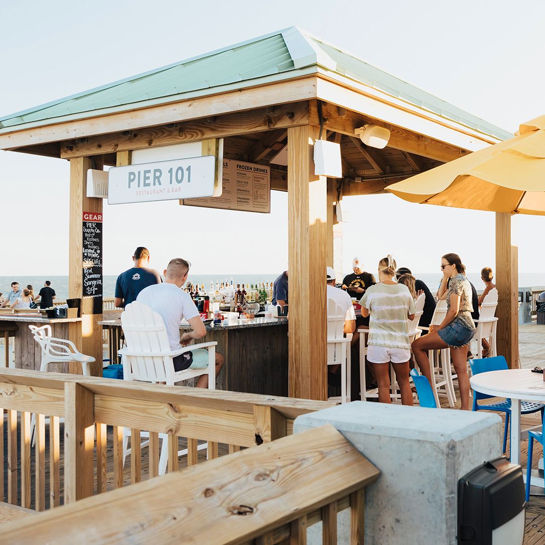 Guests gather around the open-air bar at Pier 101, enjoying drinks with ocean views in the background. The casual seating and beachside setup make it a popular spot for sunset cocktails. It captures the fun, social vibe of dining right on the pier.
