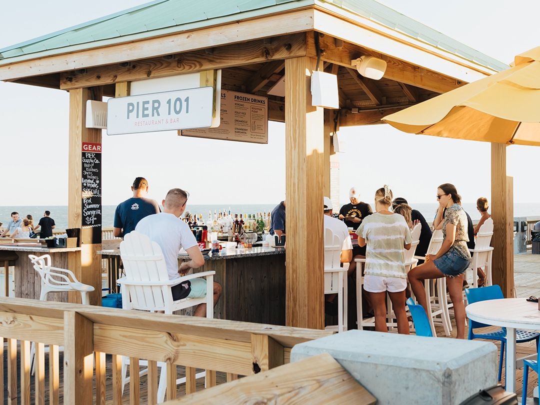 Guests gather around the open-air bar at Pier 101, enjoying drinks with ocean views in the background. The casual seating and beachside setup make it a popular spot for sunset cocktails. It captures the fun, social vibe of dining right on the pier.