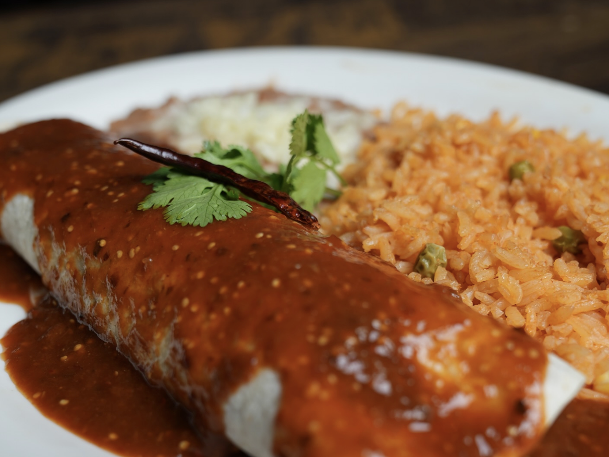 A burrito topped with rich red enchilada sauce and garnished with a dried chili and cilantro is served alongside Mexican rice and refried beans on a white plate.
