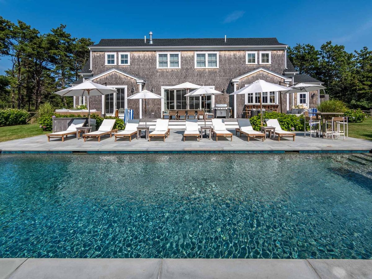 Spacious vacation home with a large private pool, surrounded by lounge chairs and umbrellas, perfect for soaking up the sun. The home features classic shingle siding and sits against a backdrop of tall pine trees, offering both luxury and privacy for family getaways or group retreats.