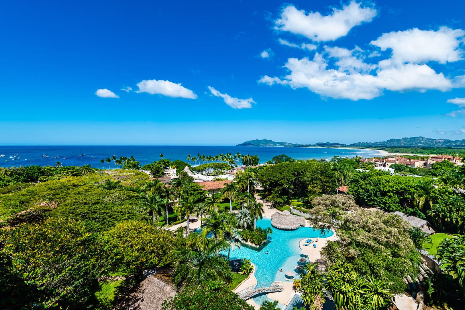 A bright oceanfront view shows the blue water, white-sand shoreline, and lush green palm trees around a large resort pool. You can see boats floating in the water and mountains in the distance. The whole scene looks sunny, tropical, and full of life.