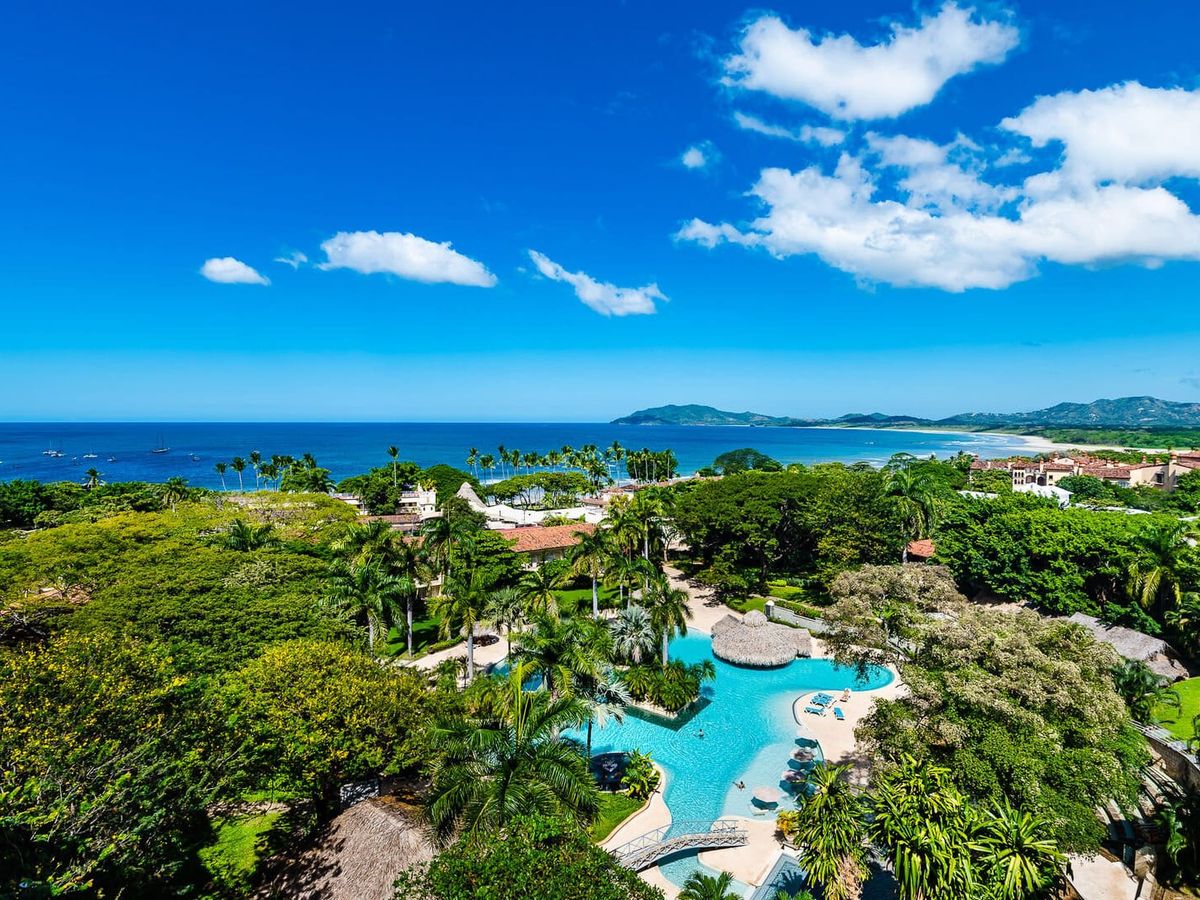 A bright oceanfront view shows the blue water, white-sand shoreline, and lush green palm trees around a large resort pool. You can see boats floating in the water and mountains in the distance. The whole scene looks sunny, tropical, and full of life.
