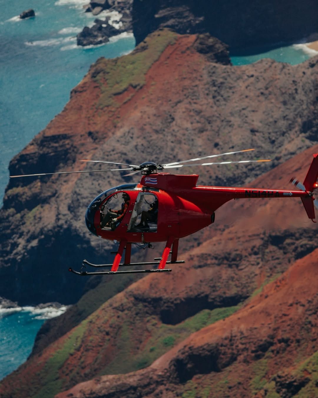 A red Airborne Aviation doors-off helicopter soars above Kauai's rugged red-clay coastline and turquoise Pacific waters during a scenic island helicopter tour.