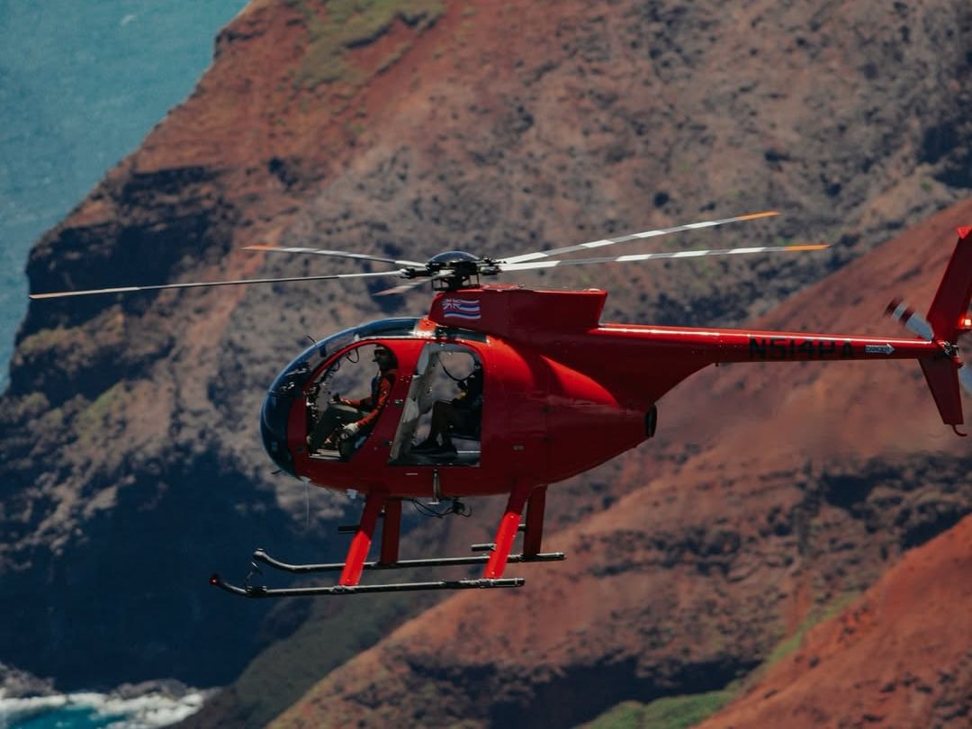 A red Airborne Aviation doors-off helicopter soars above Kauai's rugged red-clay coastline and turquoise Pacific waters during a scenic island helicopter tour.