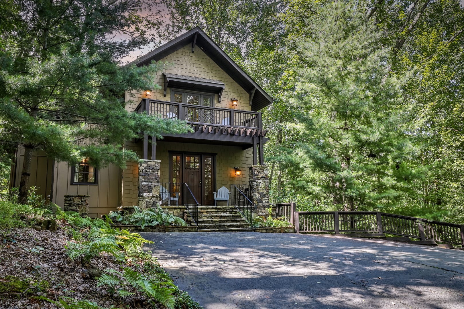 A charming two-story cabin with stone pillars, a balcony, and Adirondack chairs on the front porch, surrounded by lush green trees.