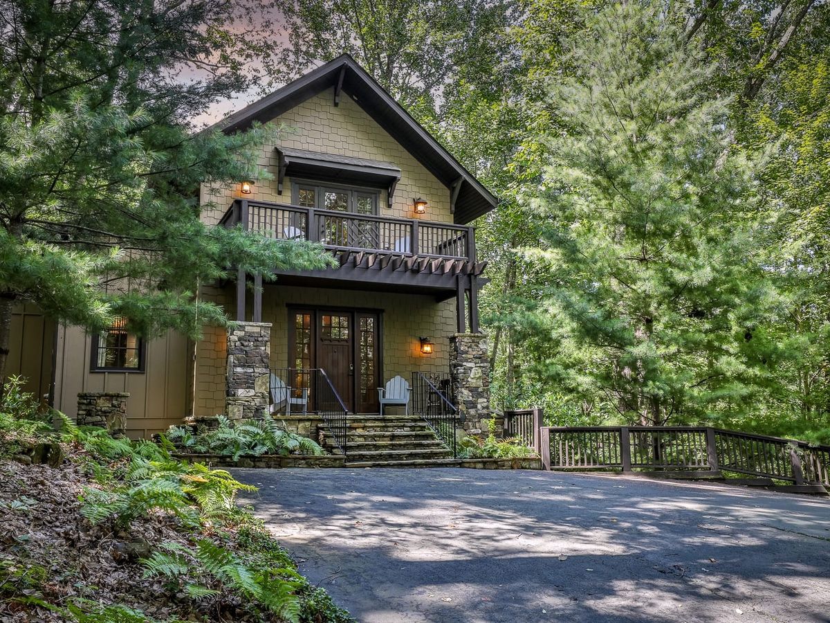 A charming two-story cabin with stone pillars, a balcony, and Adirondack chairs on the front porch, surrounded by lush green trees.