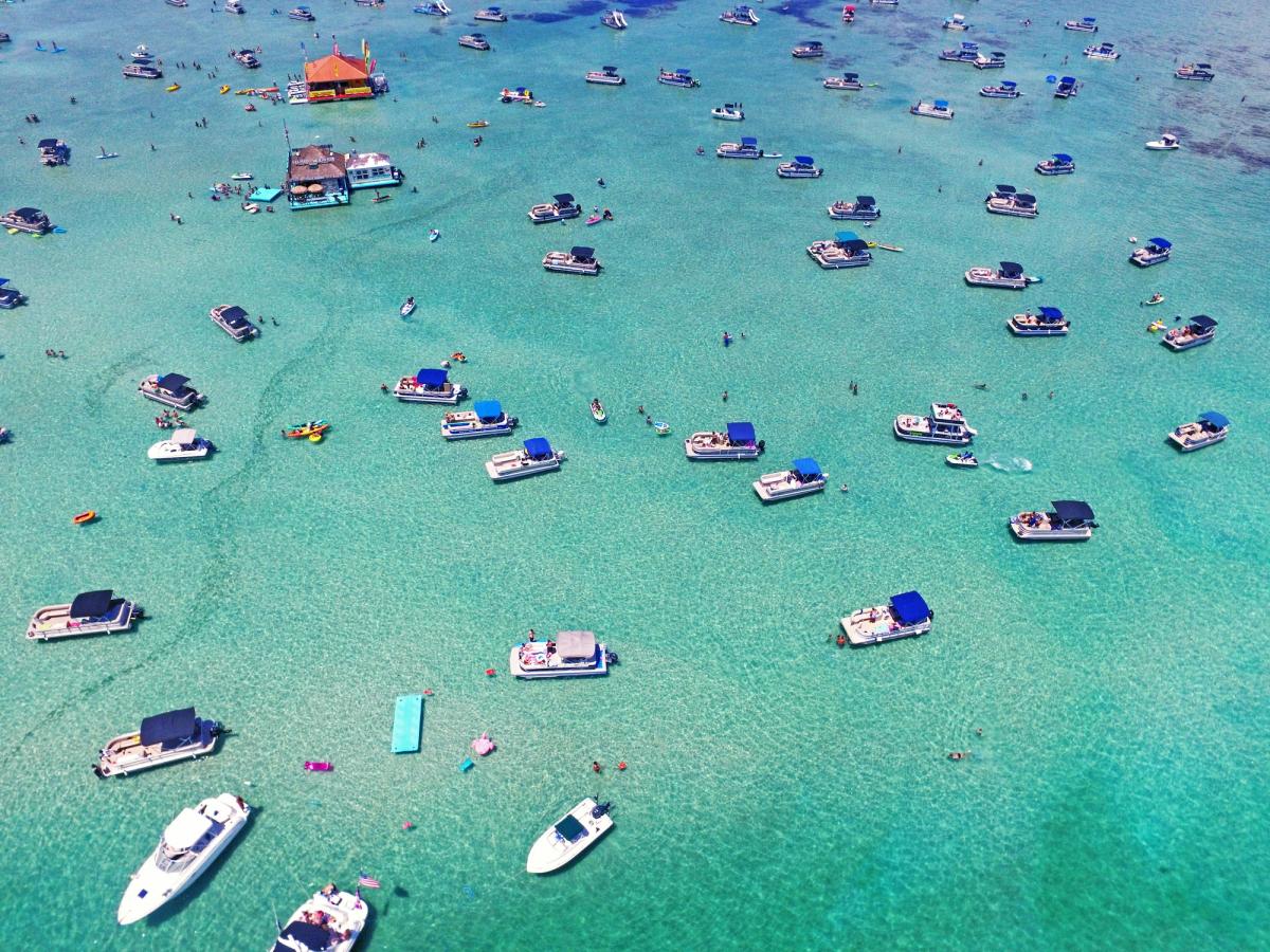 Aerial view of dozens of boats anchored in the clear turquoise waters of Crab Island in Destin, Florida, with people swimming, floating, and enjoying the vibrant sandbar party atmosphere.
