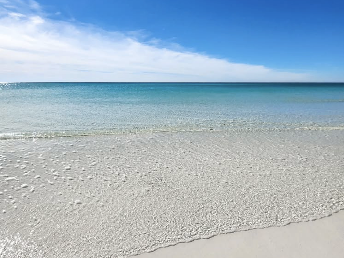 Crystal-clear turquoise water gently washing onto soft white sand under a bright blue sky with wisps of clouds on the horizon.